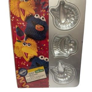 Può includere: Una teglia da forno argentata con tre stampi a forma di Big Bird e Cookie Monster di Sesame Street. La teglia è confezionata in una scatola con sfondo rosso e giallo e il testo "Big Bird & Cookie Monster Mini Pans".
