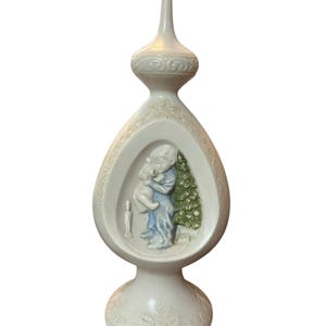 Op de afbeelding: Een witte keramische kerstboomtop met een druppelvormig midden. Het midden heeft een reliëf van een persoon die een kat vasthoudt naast een kerstboom. De topper heeft een decoratieve basis en een puntige top.