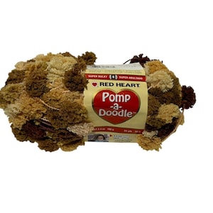 May include: A skein of Red Heart Pom-a-Doodle yarn in a brown, beige, and cream color combination. The yarn is super bulky and is labeled as "Super Bulky" and "Super Abalturado".
