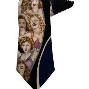 May include: A necktie featuring a repeating pattern of Marilyn Monroe portraits. The tie has a black background with a navy blue section and a white curved accent. The portraits are in shades of beige, pink, and red.