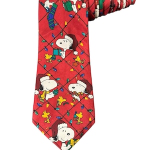 Pode incluir: Gravata vermelha com um padrão repetido de personagens Snoopy e Woodstock usando chapéus de Papai Noel e segurando presentes, com luzes de Natal. A gravata é feita de um material brilhante.