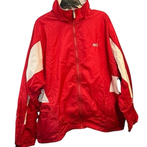 Puede incluir: Chaqueta deportiva Asics roja con paneles blancos en las mangas y hombros. Cuenta con cremallera completa y cuello alto. El logotipo de Asics es visible en el pecho.