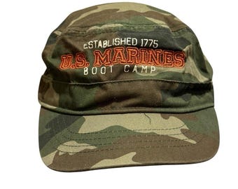 Może przedstawiać: Wojskowa czapka w kamuflażu. Tekst "ESTABLISHED 1775 U.S. MARINES BOOT CAMP" jest w kolorze pomarańczowym i beżowym. Czapka ma krótki daszek i strukturalną koronę, odpowiednią do użytku na zewnątrz.