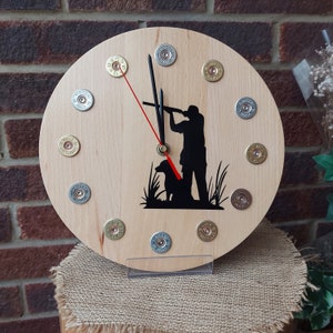 May include: A wooden clock featuring a silhouette of a hunter and a dog. The clock face is surrounded by 12 shotgun shell casings.