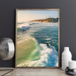 May include: A framed photograph of a surfer riding a wave in the ocean. The water is a vibrant turquoise blue and the sky is a clear blue. The surfer is wearing a wetsuit and is holding a surfboard.