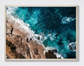 May include: An aerial view of a rocky coastline with turquoise blue ocean waves crashing on the shore. The water is a vibrant blue and green, and the rocks are a warm brown color.