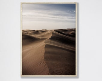 May include: A framed print featuring a desert landscape. The image showcases rolling sand dunes in shades of brown and tan under a cloudy blue sky. The print is encased in a light-colored wooden frame, adding a touch of natural warmth.