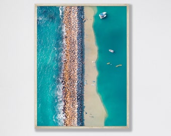 May include: An aerial view of a rocky breakwater separating turquoise blue water from a sandy beach.  The breakwater is made of large, brown rocks.  There are several small boats and kayaks in the water.