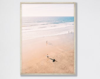 May include: An aerial view of a sandy beach with a person walking with a surfboard. The ocean is in the background with waves crashing on the shore. The sand is a light beige color.