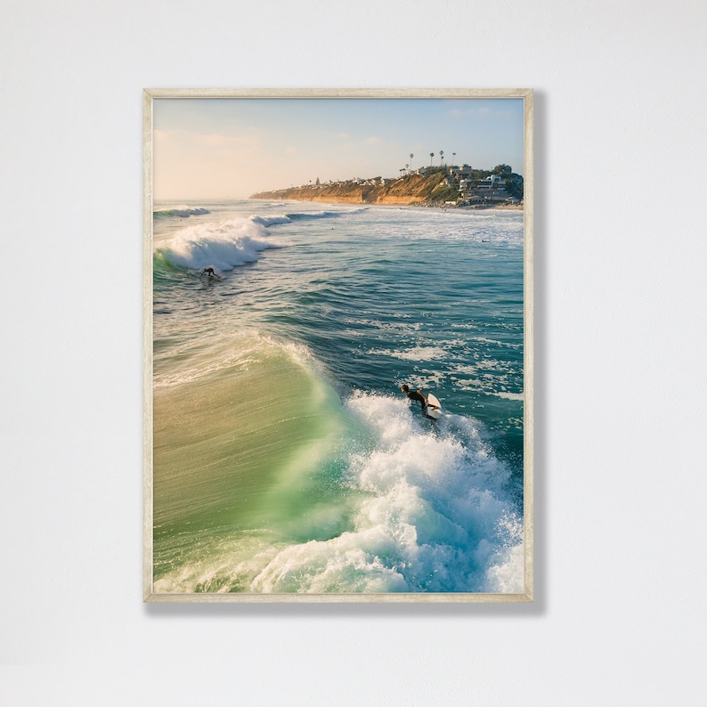 May include: A surfer rides a wave in the ocean, with a coastline and buildings in the background. The water is a vibrant blue and green, with white foam.