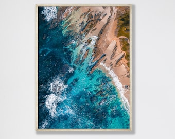 May include: An aerial view of a rocky coastline with turquoise blue water crashing against the shore. The water is clear and shallow, revealing the rocks and sand below. The beach is a light brown color and the rocks are a dark brown color.