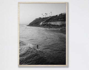 Könnte beinhalten: Schwarz-Weiß-Druck mit Rahmen, der einen Surfer auf einer Welle zeigt. Das Bild zeigt einen Surfer in Silhouette vor dem Meer, mit einer Küstenklippe und Palmen im Hintergrund. Der Druck ist in einem hellen Holzrahmen.