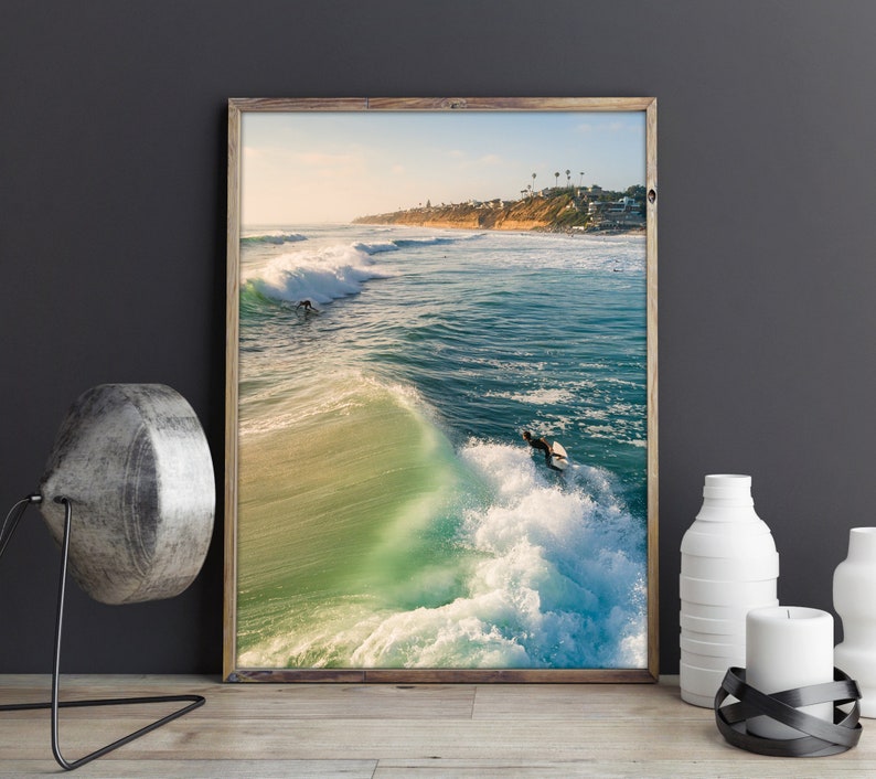 May include: A framed photograph of a surfer riding a wave in the ocean. The water is a vibrant turquoise blue and the sky is a clear blue. The surfer is wearing a wetsuit and is holding a surfboard.