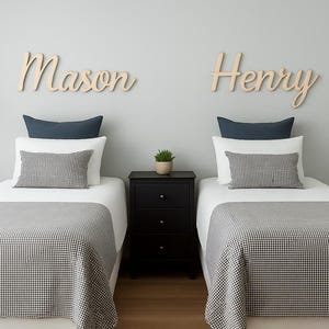 May include: Two twin beds with white bedding, gray and white checkered blankets, and decorative pillows. Wooden name signs that read "Mason" and "Henry" are mounted on the gray wall above the beds. A black nightstand sits between the beds.