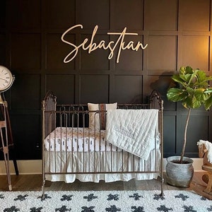 May include: A white crib with a white and black patterned sheet and a white quilt. The crib is in a room with dark brown walls and a wooden sign that says "Sebastian".