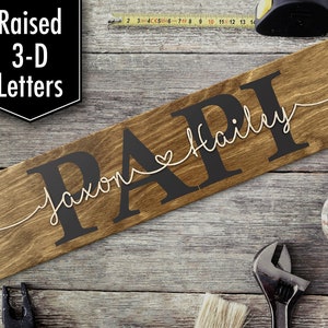 May include: A wooden sign with the word "PAPI" in large, dark letters, with "Jaxon Hailey" in a cursive font. The sign is brown and has a rustic look. The words "Raised 3-D Letters" are in a black box in the upper left corner.