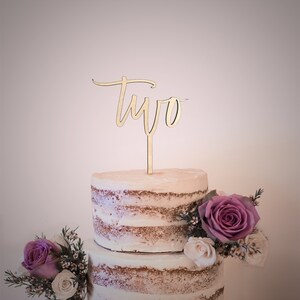 May include: A two-tiered cake decorated with flowers and a wooden cake topper that says "two" in cursive.