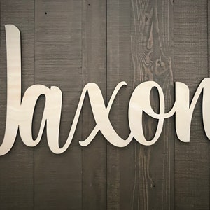 May include: White wooden cutout of the name "Jaxon" in a cursive font, mounted on a dark wood background.