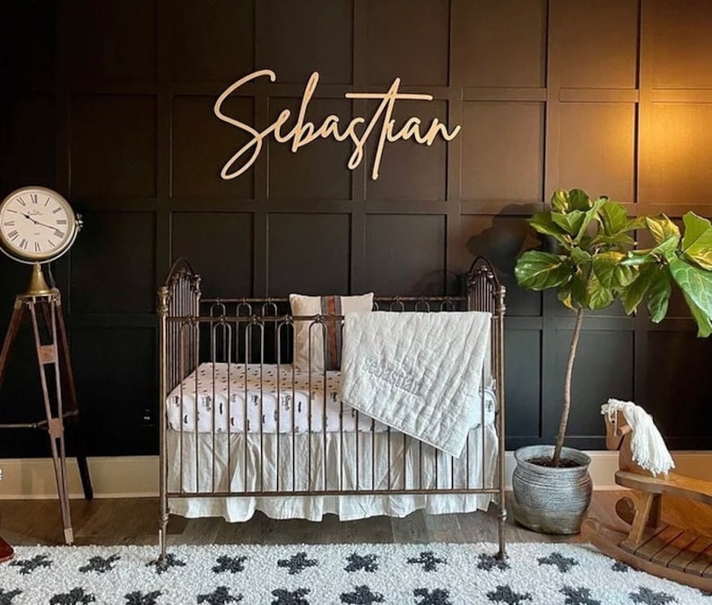 May include: A white crib with a white and black patterned sheet and a white quilt. The crib is in a room with dark brown walls and a wooden sign that says "Sebastian".