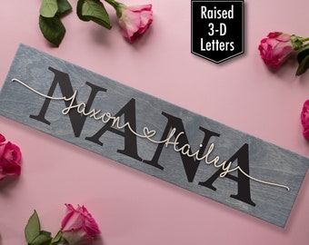 May include: A wooden sign with the word "NANA" in large, raised, black letters. The names "Jaxon" and "Hailey" are written in white string above and below the word "NANA" with a small heart between the names. The sign is on a pink background with pink roses.