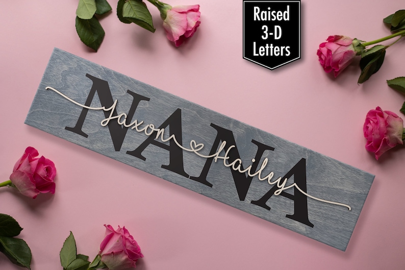 Op de afbeelding: Een houten bord met het woord "NANA" in grote, reli&euml;f, zwarte letters. De namen "Jaxon" en "Hailey" zijn geschreven in wit touw boven en onder het woord "NANA" met een klein hartje tussen de namen. Het bord is op een roze achtergrond met roze rozen.