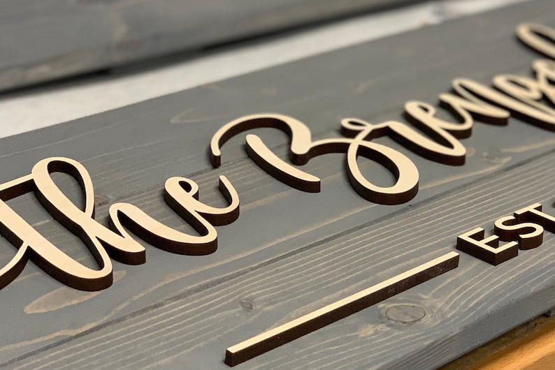 Puede incluir: Un letrero de madera con las palabras "The Brown" en escritura cursiva y "EST" debajo. El letrero est&aacute; pintado de color gris oscuro y las letras son de color madera claro.
