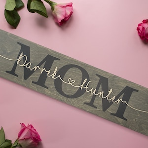 May include: A wooden sign with the word "MOM" in large black letters. The names "Derek" and "Hunter" are written in cursive above and below the word "MOM" with a small heart between the names.