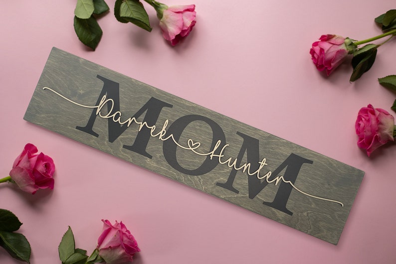 May include: A wooden sign with the word "MOM" in large black letters. The names "Derek" and "Hunter" are written in cursive above and below the word "MOM" with a small heart between the names.