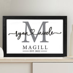 May include: A framed print with a gray letter M, the names "ryan" and "nicole" written in cursive with a heart symbol between them, and the text "MAGILL EST. 2022" below.