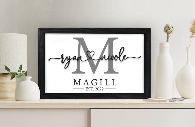 May include: A framed print with a gray letter M, the names "ryan" and "nicole" written in cursive with a heart symbol between them, and the text "MAGILL EST. 2022" below.