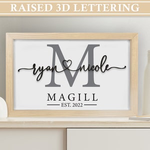 May include: A wooden framed print with a gray letter M, the names "ryan" and "nicole" written in cursive with a heart symbol between them, and the text "MAGILL EST. 2022" below.