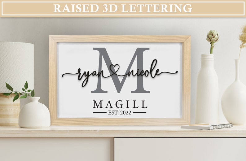 May include: A wooden framed print with a gray letter M, the names "ryan" and "nicole" written in cursive with a heart symbol between them, and the text "MAGILL EST. 2022" below.
