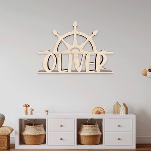 May include: Wooden name sign with a ship's wheel design and the name "Oliver" in a split design. The sign is mounted on a white wall above a white dresser with two baskets and a stuffed giraffe.