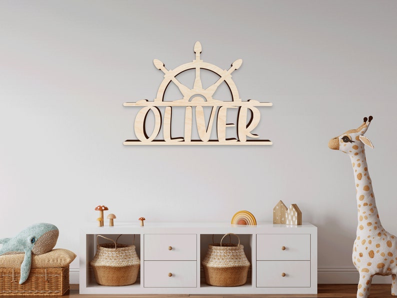 May include: Wooden name sign with a ship's wheel design and the name "Oliver" in a split design. The sign is mounted on a white wall above a white dresser with two baskets and a stuffed giraffe.