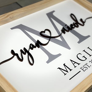 May include: A framed wooden sign with the last name "MAGILL" and the text "EST. 2022" in black lettering. The names "Ryan" and "Nicole" are written in cursive with a heart symbol between them. The letter "M" is in gray and the names are in black.