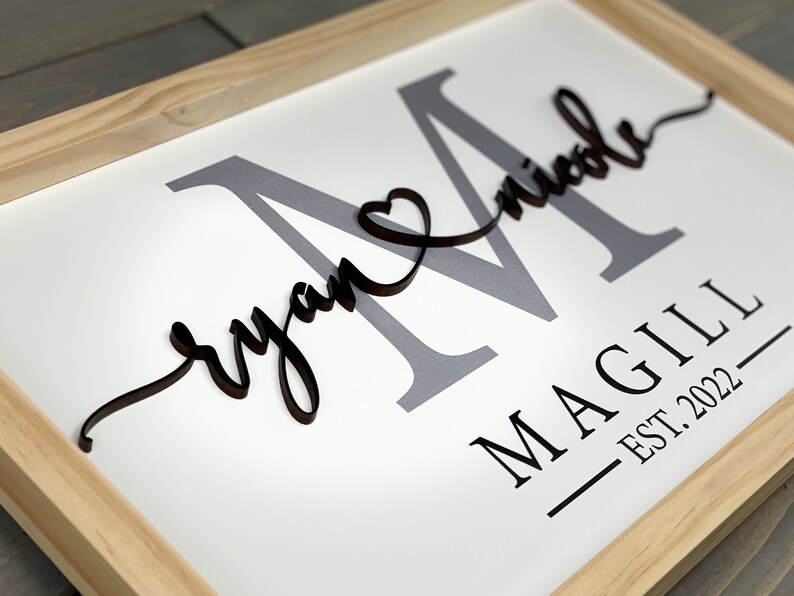 May include: A framed wooden sign with the last name "MAGILL" and the text "EST. 2022" in black lettering. The names "Ryan" and "Nicole" are written in cursive with a heart symbol between them. The letter "M" is in gray and the names are in black.