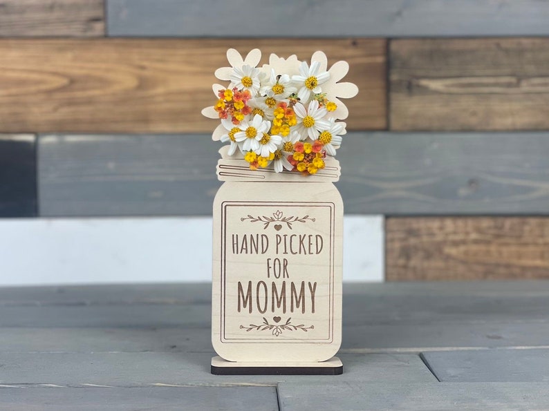 May include: Wooden cutout of a mason jar with a bouquet of white and yellow flowers. The jar has the text "HAND PICKED FOR MOMMY" on it.