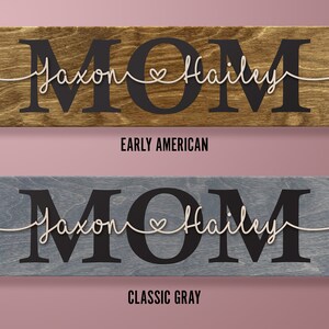 May include: Wooden sign with the word "MOM" in large black letters. The names "Jaxon" and "Hailey" are written in cursive with a heart between them. The sign is available in two colors: Early American and Classic Gray.