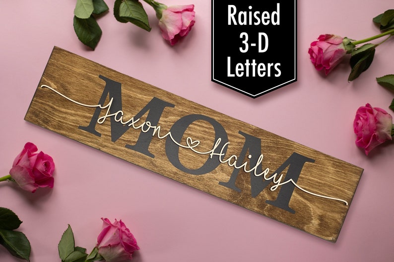 May include: A wooden sign with the word "MOM" in raised black letters. The names "Jaxon" and "Hailey" are written in white cursive script on either side of the word "MOM" with a small heart between the names.