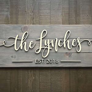 Puede incluir: Un letrero de madera gris con el texto "the Lynches" en letras blancas cursivas y "EST. 2018" en letras blancas debajo.