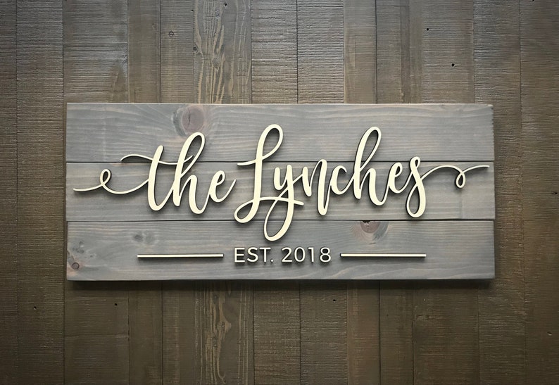 Puede incluir: Un letrero de madera gris con el texto "the Lynches" en letras blancas cursivas y "EST. 2018" en letras blancas debajo.