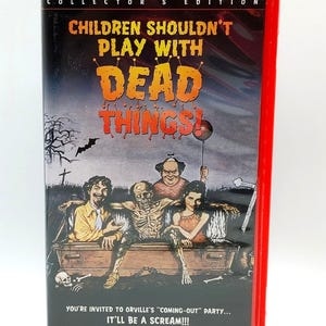 Peut inclure: Un boîtier VHS rouge pour l'édition collector de Children Shouldn't Play With Dead Things. La couverture montre une scène de cimetière avec un squelette dans un cercueil, deux personnes et un homme tenant un ballon. Le texte inclut le titre.