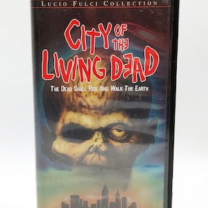 May include: A black VHS case for the Lucio Fulci Collection film City of the Living Dead. The cover features a large, decaying zombie head above a city skyline silhouette, with the text City of the Living Dead and The Dead Shall Rise And Walk The Earth.