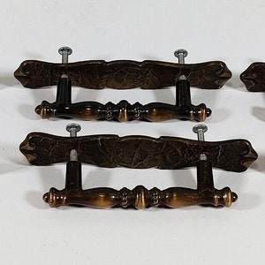 以下が含まれることがあります： 装飾的なデザインが施されたアンティークブラス製のキャビネットドアプル8個セット。各プルは、中央のバーと2本のネジが付いた装飾的な曲線形状をしています。