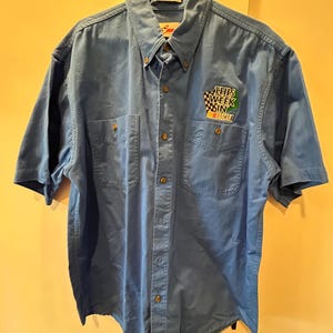 Puede incluir: Camisa azul claro de manga corta con dos bolsillos en el pecho y botones. El texto "THIS WEEK IN NASCAR" está en el pecho derecho. La camisa está colgada de una percha metálica.