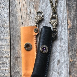 May include: Two leather cases, one tan and one black, with decorative metal buttons and stitching. The cases have metal clasps and are attached to a chain. The tan case has a circular button, while the black case has a button with purple accents.