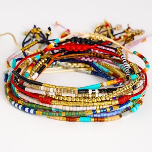 May include: A stack of colorful beaded bracelets. The bracelets feature a variety of colors including gold, red, blue, and turquoise. The beads are small and strung together on a cord. Some bracelets have adjustable knots.