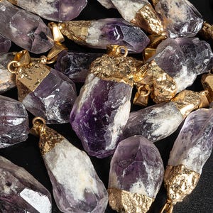 Könnte beinhalten: Eine Sammlung von Amethyst-Kristallanhängern mit vergoldeten Einstellungen. Die Kristalle sind in verschiedenen Formen und Größen, einige haben eine klare, fast durchscheinende Erscheinung, während andere ein tiefes Lila haben.