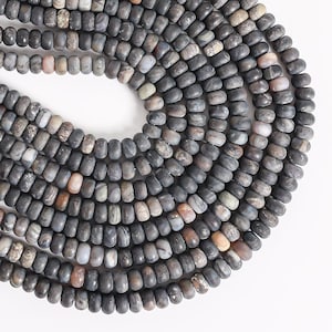 May include: A collection of round, disc-shaped beads arranged in a curved pattern. The beads feature a mottled texture with a color palette of charcoal gray, slate, and muted earthy tones, including subtle hints of beige and soft pink.