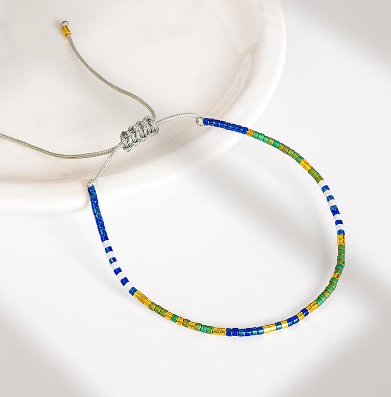 May include: Adjustable beaded bracelet featuring a colorful pattern of blue, green, gold, and white beads. The bracelet has a silver-colored cord and a sliding knot closure for a customizable fit. This piece is a stylish accessory.
